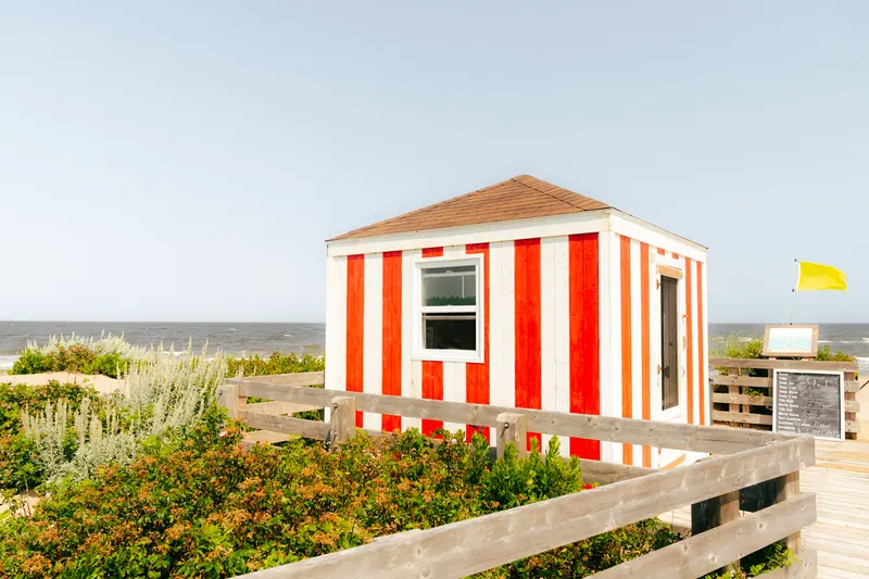 A red and white striped beach hut behind dune grass and a wooden boardwalk, with the ocean beyond.