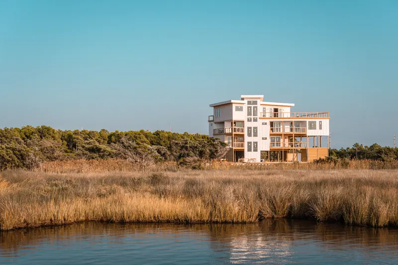 A white multi-storey house standing alone across reeds and still water in warm evening light.