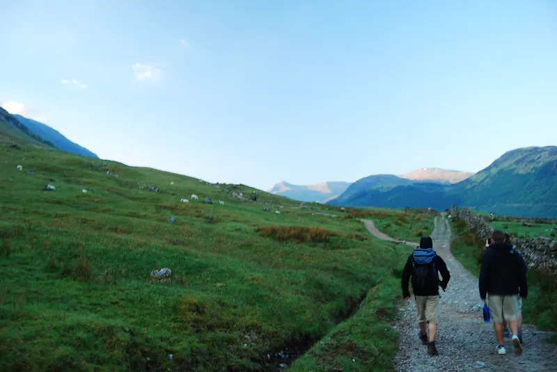 Two hikers walking away from the camera along a gravel path through green hills toward distant mountains.