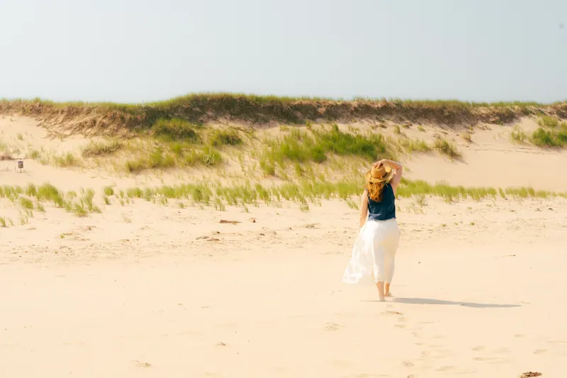 A woman in a straw hat and white skirt walking across a sandy beach toward grassy dunes.