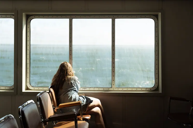 A woman sitting inside a boat cabin, looking out a large window at the open sea.