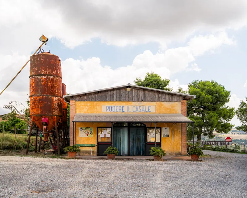 A yellow country shop with a rusty grain silo beside it and pine trees behind, under a sign reading Podere il Casale.