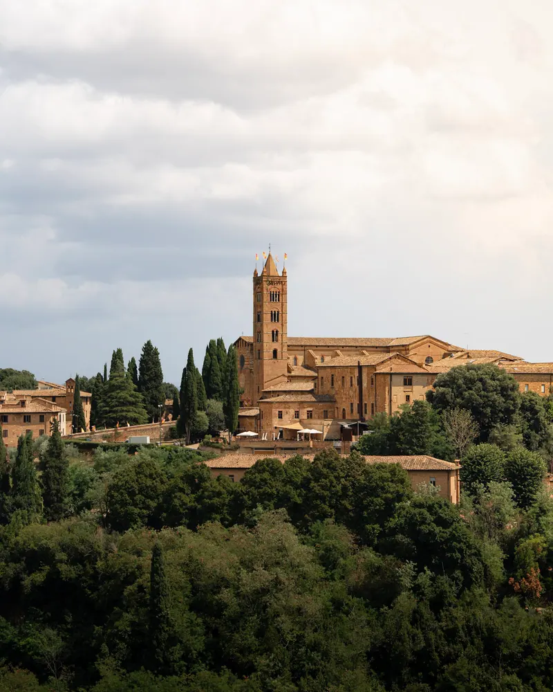 A stone church with a tall bell tower rising above green trees on a hillside under cloudy skies.